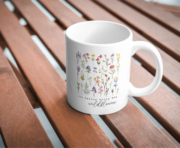 Weiße Keramiktasse mit zarten Aquarell-Wildblumen in verschiedenen Farben und dem Zitat „You belong among the wildflowers“, fotografiert auf einem Holztisch.